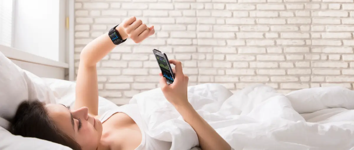 An image of a woman looking at her smartwatch and phone while laying in bed